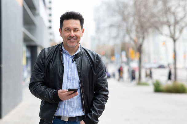 Muotokuva komea kypsä latino mies yllään nahkatakki älypuhelin seisoo kaupungin ulkopuolella - Valokuva, kuva