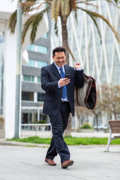 Πλήρες πορτρέτο του ευτυχισμένου ώριμου επιχειρηματία που περπατά στην πόλη με την τσάντα ταξιδιού κοστούμι και χρησιμοποιώντας το κινητό του τηλέφωνο - Φωτογραφία, εικόνα