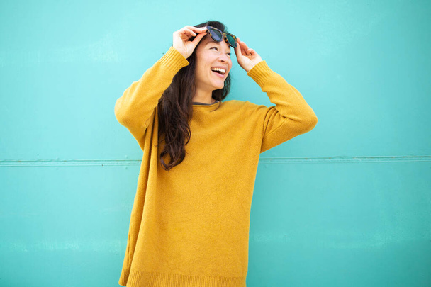 Portret van een mooie jonge vrouw met een zonnebril die wegkijkt en glimlacht tegen de groene muur - Foto, afbeelding