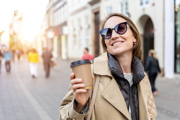 Портрет молодої красивої щасливої усміхненої жінки, яка п'є каву, щоб піти на чай в екологічно стійкій паперовій чашці, одягненій в траншею, насолоджується прогулянкою по європейській вулиці міста. Стильна жінка міський спосіб життя. - Фото, зображення