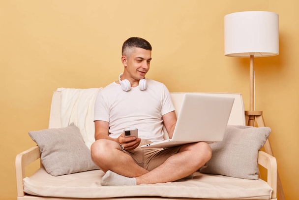 Internet browsing. Home office. Leisure surfing. Positive attractive young man working on laptop computer and using smartphone sitting on comfortable couch at home. - Foto, imagen
