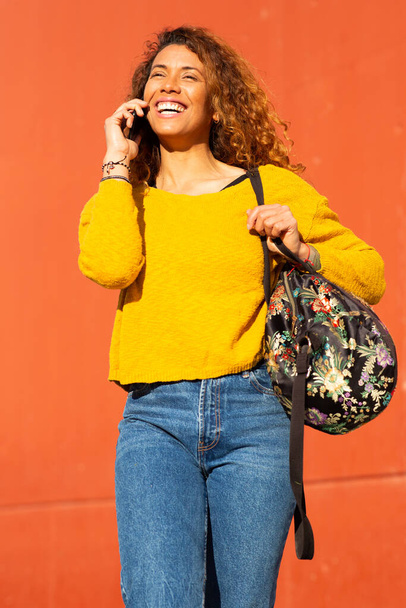 Portrait of beautiful young latin woman talking on mobile phone while walking against orange wall - 写真・画像
