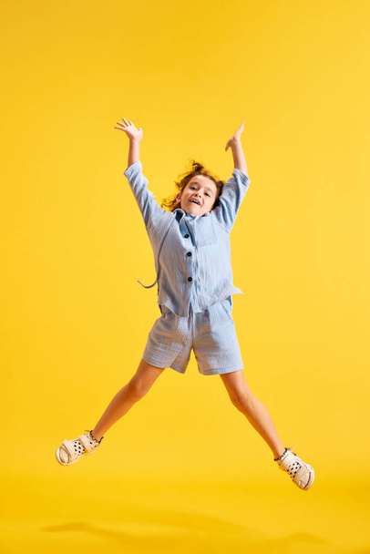 Full-length portrait of positve, active little girl, child in casual clothes cheerfully jumping against yellow studio background. Concept of emotions, childhood, education, fashion, lifestyle, ad - Fotoğraf, Görsel