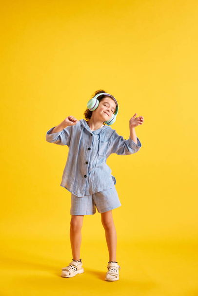 Full-length portrait of beautiful little child, girl listening to music in headphones against yellow studio background. Concept of emotions, childhood, education, fashion, lifestyle, ad - Φωτογραφία, εικόνα