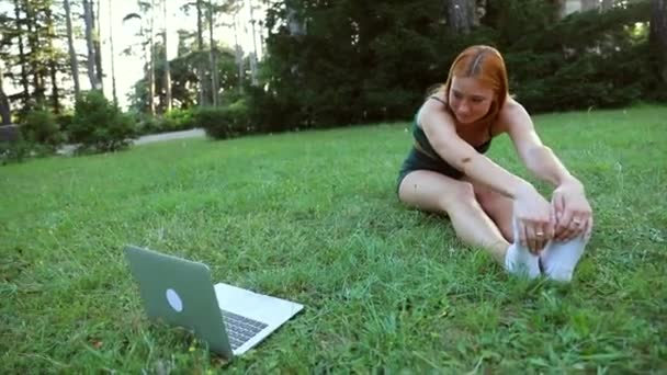 Девушка растягивается во время занятий онлайн на своем ноутбуке в общественном парке - Кадры, видео