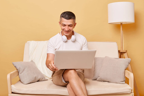Digital leisure. Notebook work. Internet browsing. Freelancer connection. Cheerful joyful young man working on laptop computer sitting on comfortable sofa at home. - 写真・画像