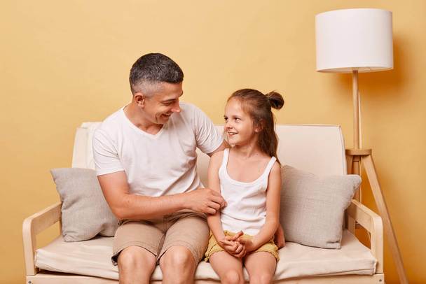 Positive friendly father talking with his daughter sitting on cough at home against beige wall spending good time together enjoying moment. - Foto, Bild