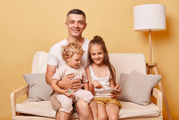Friendly delighted happy father sitting on cozy sofa couch using smartphone with little girls daughters at home against beige wall spending time together surfing internet. - Фото, изображение