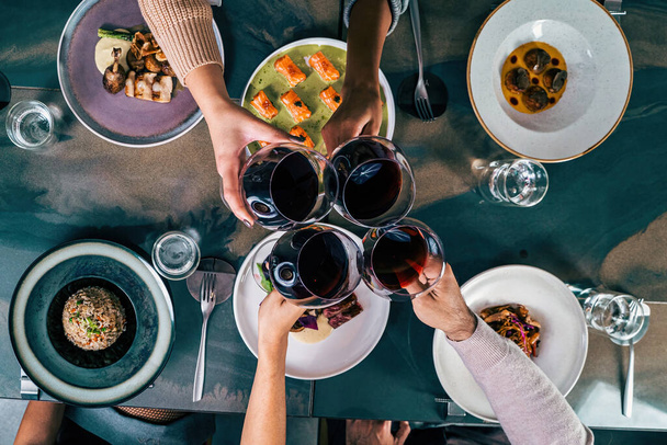 Vista aérea de cuatro amigos haciendo un brindis con vino tinto sobre una mesa de restaurante de fusión con varios platos - Foto, imagen