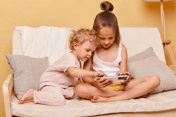 Concentrated children little girls sisters sitting on couch using smartphone together posing against beige wall playing on mobile phone looking at device display. - Photo, image