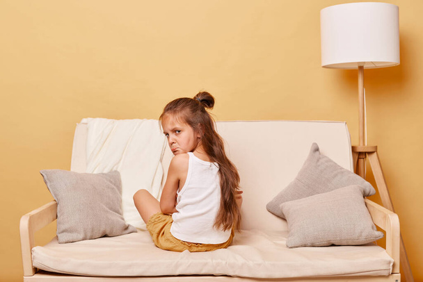 Upset little girl sitting backwards on couch at home, depressed female child having bad mood, offended kid feeling sadness, turning and looking at camera. - Photo, Image