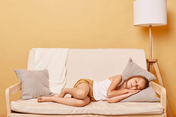 Full length of sleeping little girl lying on a couch with hand under cheek keeps eyes closed resting at home day napping relaxing during free time. - Foto, imagen