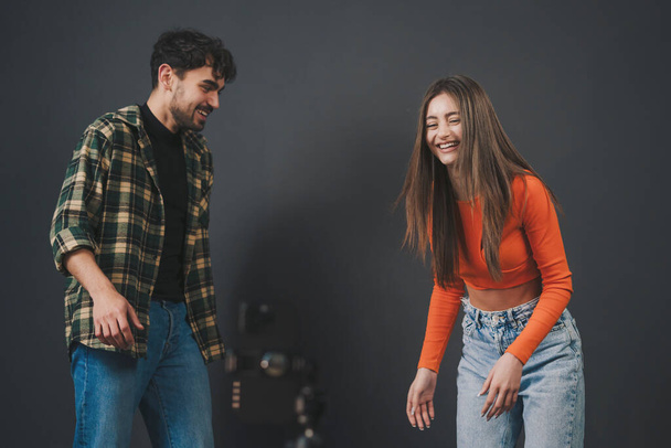 Happy young man and woman in casual clothes smiling and looking at camera while standing against black background - Photo, Image