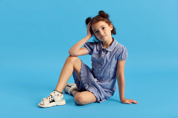 Portrait of little girl, child with cury hair in cute dress sitting on floor and posing against blue studio background. Concept of emotions, childhood, education, fashion, lifestyle, ad - Foto, Imagen