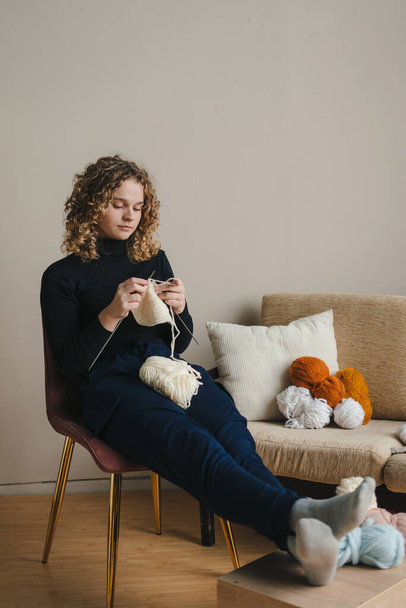  женщина с вьющимися волосами улыбается и смотрит в камеру, сидя на диване и плетя шарф - Фото, изображение