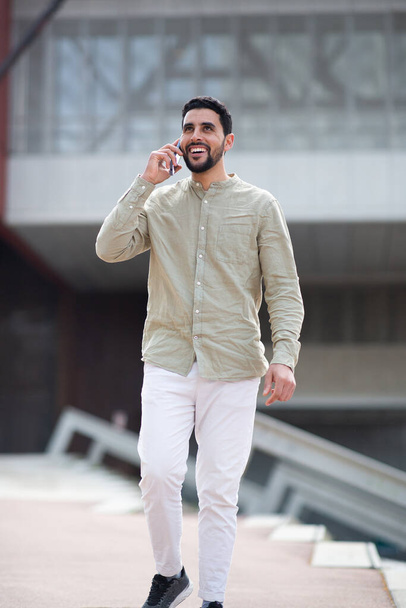 Πορτρέτο του όμορφου νεαρού αραβικού άνδρα με τα πόδια και μιλώντας στο κινητό τηλέφωνο έξω - Φωτογραφία, εικόνα