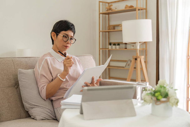Впевнена літня бізнес-леді робить паперові роботи, сидячи з планшетом на дивані вдома. - Фото, зображення
