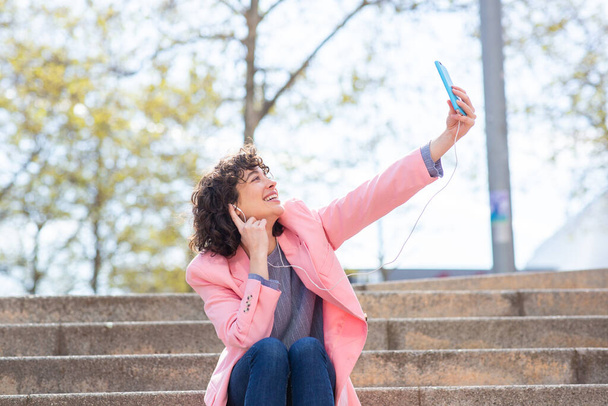 Portrait of attractive woman wearing earphones sitting on city steps and taking selfie with her mobile phone - Foto, Imagem