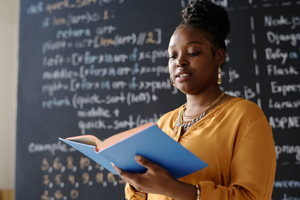 African American teacher reading book at IT lesson standing against blackboard - Foto, imagen