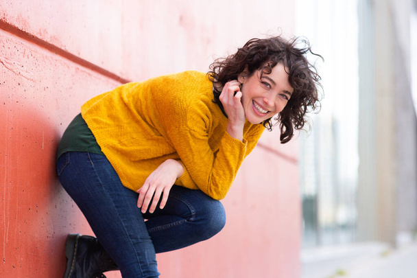 Portrait of attractive young woman bending over looking at camera and smiling by the wall outdoors - Photo, Image
