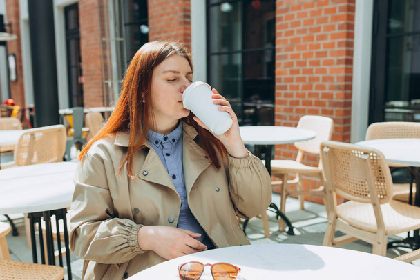 Vrolijke vrolijke jonge vrouw in mode kantoorkleding en zonnebrandcrème met eco kopje thee. Mockup, vrouw met bio koffie papier beker en zitten op het terras van het cafe op de straat van de stad. - Foto, afbeelding