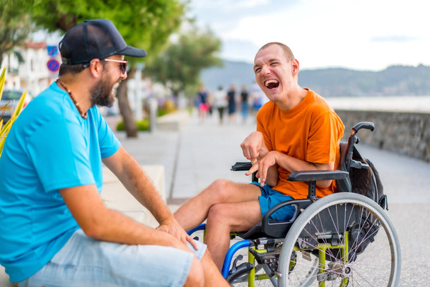A disabled person in a wheelchair with a friend on summer vacation having fun laughing a lot by the sea - Zdjęcie, obraz