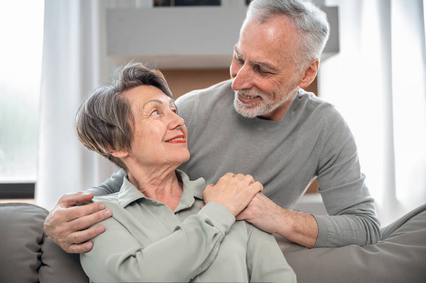 Happy mature couple looking to each other and hugging. Elderly man care about his smiling wife while she sitting on comfort sofa at living room. People enjoying tender moment together - Photo, Image