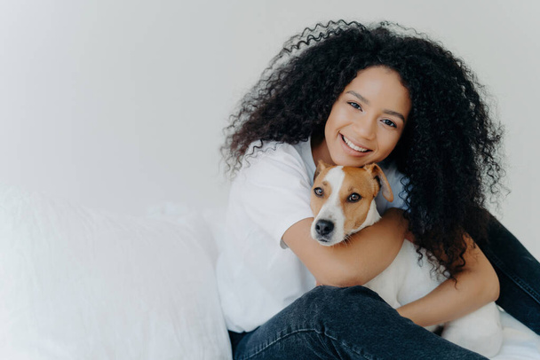 Mujer joven con Afro, abraza perro, cuida, sonríe, ropa casual, fondo blanco, cama, espacio de promoción.  - Foto, Imagen