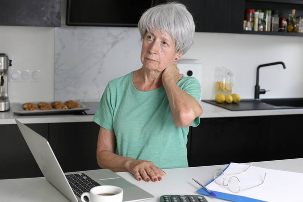 close-up portret van volwassen vrouw met nekpijn tijdens het werken met laptop in de keuken - Foto, afbeelding