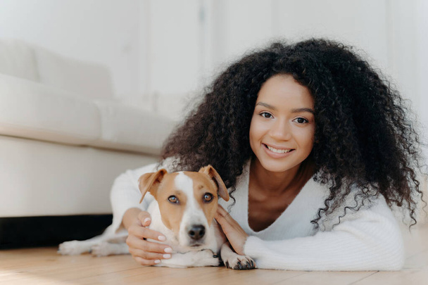 Όμορφη κοπέλα με μαλλιά Afro βρίσκεται στο πάτωμα του σαλονιού, μοιράζονται ευχάριστα συναισθήματα με πρόσφατα υιοθετημένο σκυλί. Αγαπημένο κατοικίδιο σε νέο διαμέρισμα, δημιουργώντας όμορφες στιγμές μαζί. - Φωτογραφία, εικόνα