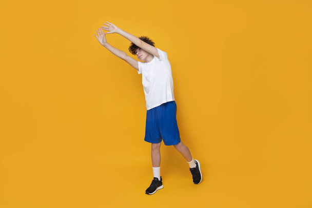 Ένας έφηβος με αθλητική στολή κάνει προθέρμανση σε κίτρινο φόντο. - Φωτογραφία, εικόνα