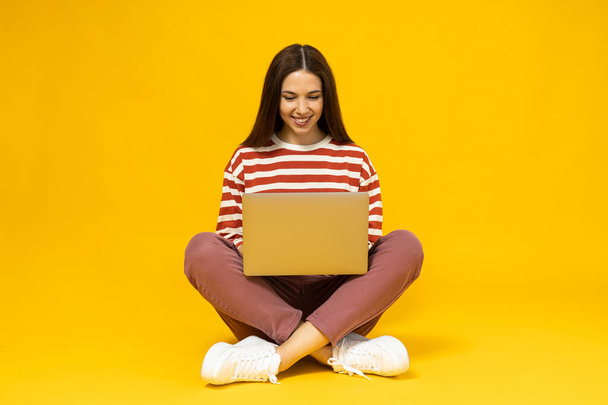 Una joven con una camiseta a rayas se sienta con un portátil sobre un fondo amarillo - Foto, Imagen