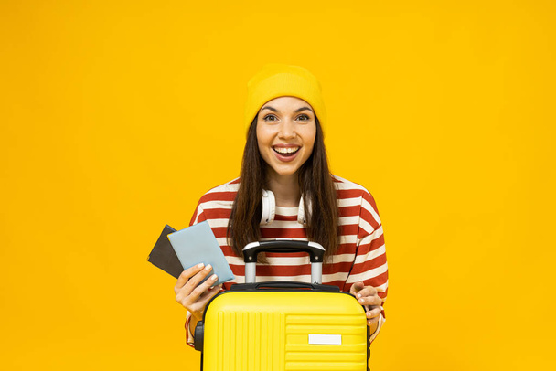 Happy young woman in striped t-shirt with suitcase and tickets on yellow background - Photo, Image