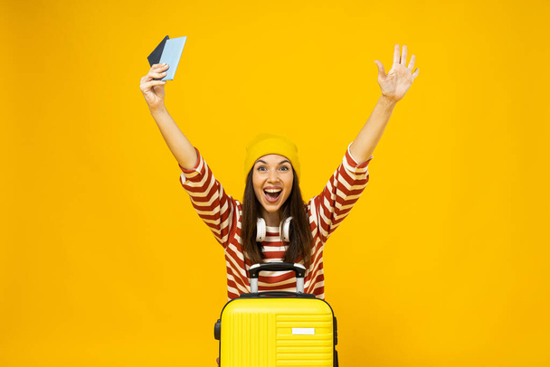 Happy young woman in striped t-shirt with suitcase and tickets on yellow background - Photo, image