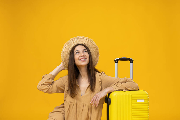 Νεαρή γυναίκα με καπέλο παραλίας και βαλίτσα σε κίτρινο φόντο - Φωτογραφία, εικόνα