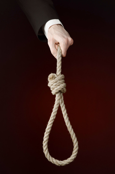 Suicide and business topic: Hand of a businessman in a black jacket holding a loop of rope for hanging on dark red isolated background - 写真・画像