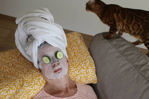 primer plano retrato de mujer madura relajante con toalla en la cabeza, máscara facial y pepinos en los ojos - Foto, Imagen
