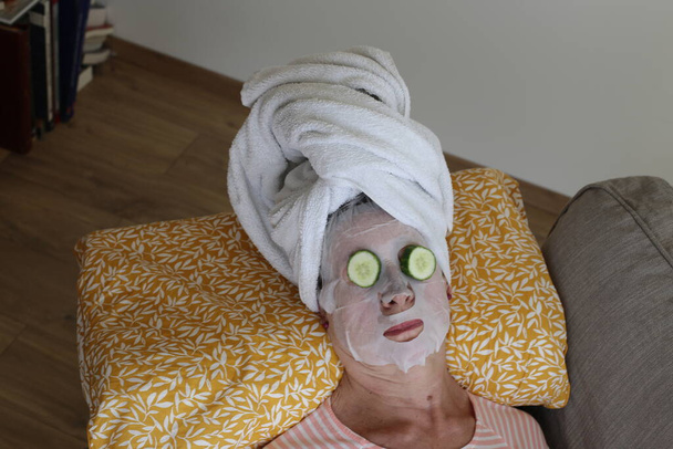 primer plano retrato de mujer madura relajante con toalla en la cabeza, máscara facial y pepinos en los ojos - Foto, Imagen