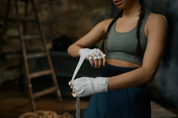 Kadın boksörün eşofman giyip ellerini bandajla sarmalayıp eski spor kulübünün boks çantasının yanında durduğu yakın plan. - Fotoğraf, Görsel