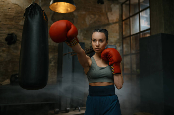 Πορτρέτο της νεαρής γυναίκας πυγμάχος εξάσκηση γροθιές φορώντας γάντια πυγμαχίας αναζητούν συμπυκνωμένο και με αυτοπεποίθηση - Φωτογραφία, εικόνα