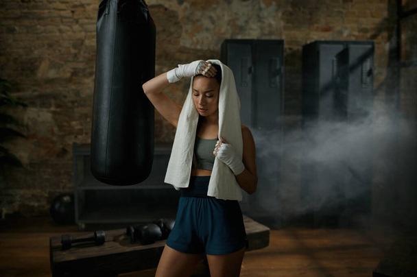zmęczona sportowa atrakcyjna kobieta bokser noszenie ręcznik na głowie i kickboxing bandaże stojąc na siłowni zrobić przerwę po ciężkim treningu - Zdjęcie, obraz
