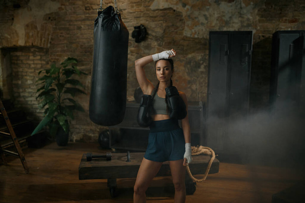 Serieuze vrouw bokser poseren voor camera gekleed in sportkleding, handen verband dragen bokshandschoenen op schouders staande over zolder sportschool interieur - Foto, afbeelding