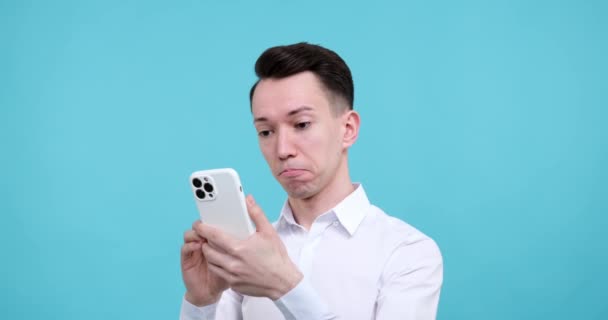 A Caucasian man standing against a vibrant blue background. With a sense of awe in his eyes, he holds a smartphone in his hands, embarking on a captivating digital journey. - Footage, Video