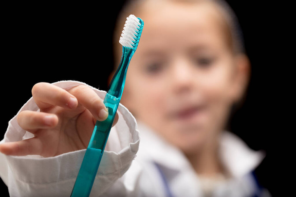 Mosd meg a fogad! Mondja a kislány fogorvos. Egy kicsi, elmosódott lány túlméretezett kabátban fogorvost játszik, bemutatva egy fogkefét (fókuszban), arra sürgetve Önt, hogy mosson fogat az egészsége érdekében, és hogy elkerülje a rossz leheletet. - Fotó, kép