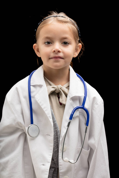 Small girl plays doctor, half-body portrait on black background. She wears an oversized white coat and stethoscope. This cute blonde girl might prefer to become a vet when she grows up - Photo, Image