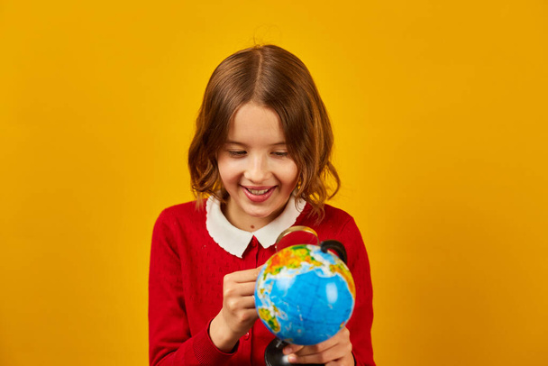Stylish smiling school teenage girl holding magnifying glass in her hand and look on globe on yellow studio background, back to school concept - Foto, Imagem