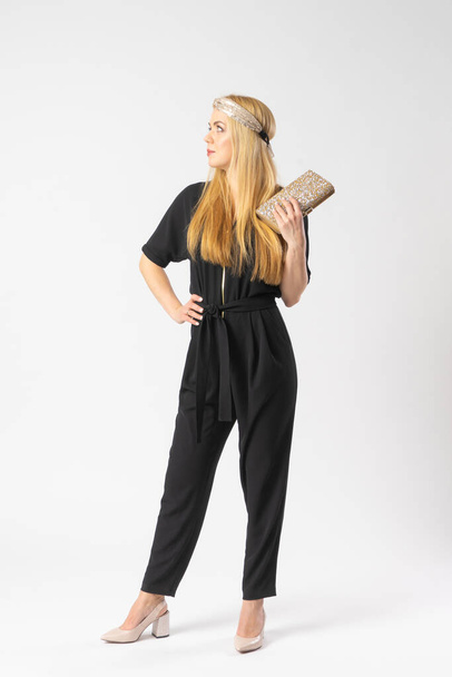 A young blonde woman in a smart black trouser suit with a golden clutch bag in her hand. Model posing in the studio on a white background. - Фото, изображение