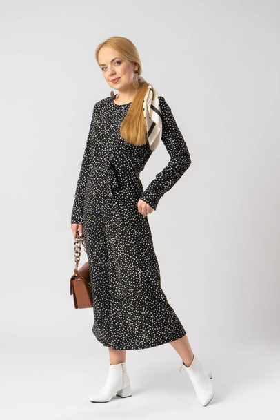 A young blonde woman in an elegant casual black trouser suit with white polka dots, with a small brown leather handbag. Model posing in the studio on a white background. - Photo, image