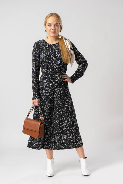 A young blonde woman in an elegant casual black trouser suit with white polka dots, with a small brown leather handbag. Model posing in the studio on a white background. - Fotoğraf, Görsel