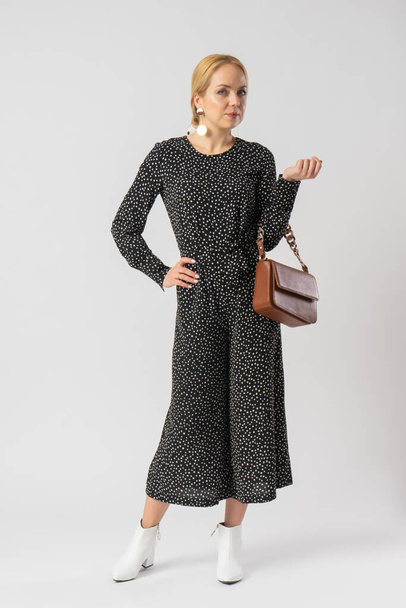 A young blonde woman in an elegant casual black trouser suit with white polka dots, with a small brown leather handbag. Model posing in the studio on a white background. - Foto, immagini